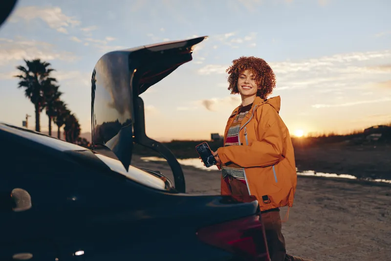 Woman lifestyle portrait by car hood open on road at sunset, curly hair, orange jacket, smiling with smartphone and travel bag, checking vehicle for trip, warm golden cinematic light.