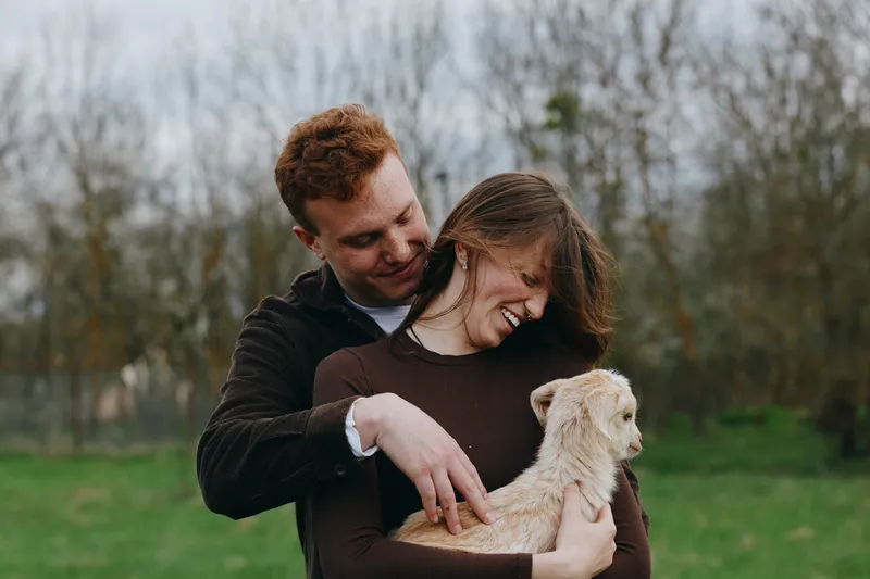 Bright candid scene shows a man and woman embracing outdoors with a small dog in a park, capturing warmth, joy, and affectionate connection amid open grass and trees