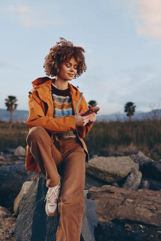 Woman lifestyle outdoors, curly hair, wearing brown jacket and overalls, sitting on rocks, looking at phone, relaxed travel mood at sunset, autumn countryside, peaceful portrait