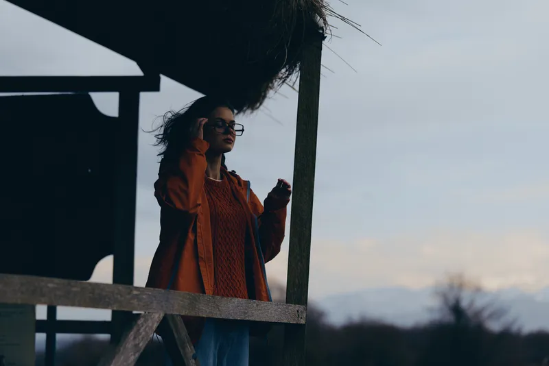 Woman in lifestyle outdoors scene, standing by wooden fence, profile view, wearing orange coat, wind moving hair, sunset evening light, cloudy sky, calm mood, soft breeze, relaxed posture