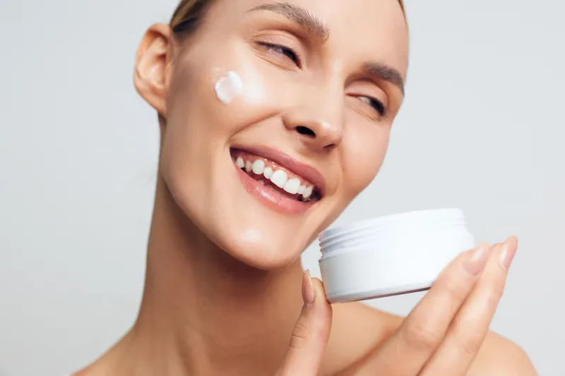Woman face cream skincare beauty closeup, smiling female applies moisturizer from a white jar, smooth skin glow on neutral studio background, enjoying relaxed self care moment today.