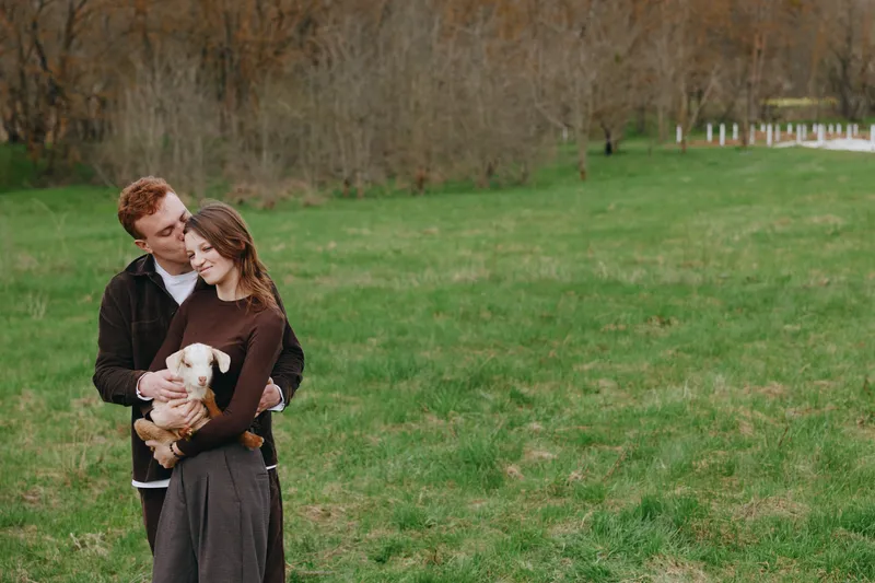 A couple smiles in a sunlit park, one embracing a small dog. The scene captures warmth, companionship, and everyday romance in a bright, open green field.