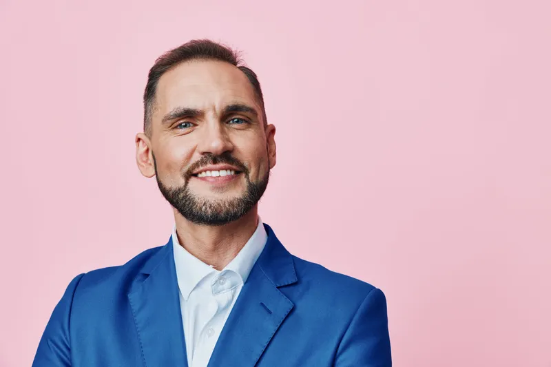Professional man in a blue suit on a solid pink background, portraying confident and friendly emotions. Isolated color backdrop enhances a modern business portrait.