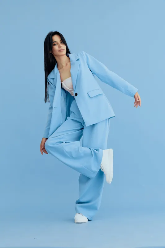 Woman in blue suit posing gracefully against pastel blue background, wearing white sneakers and light inner blouse, showcasing elegant contemporary fashion style.