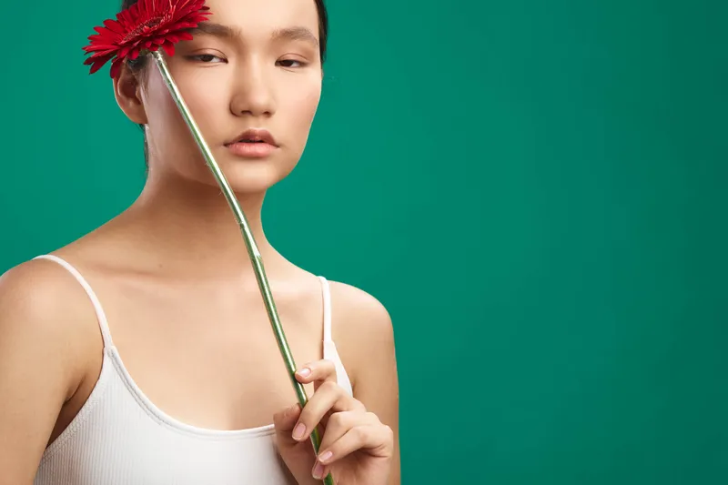 asian woman holds a vibrant flower near her face against a solid green backdrop, creating a serene portrait with soft lighting and minimal styling, complemented by warm tones, subtle texture, and