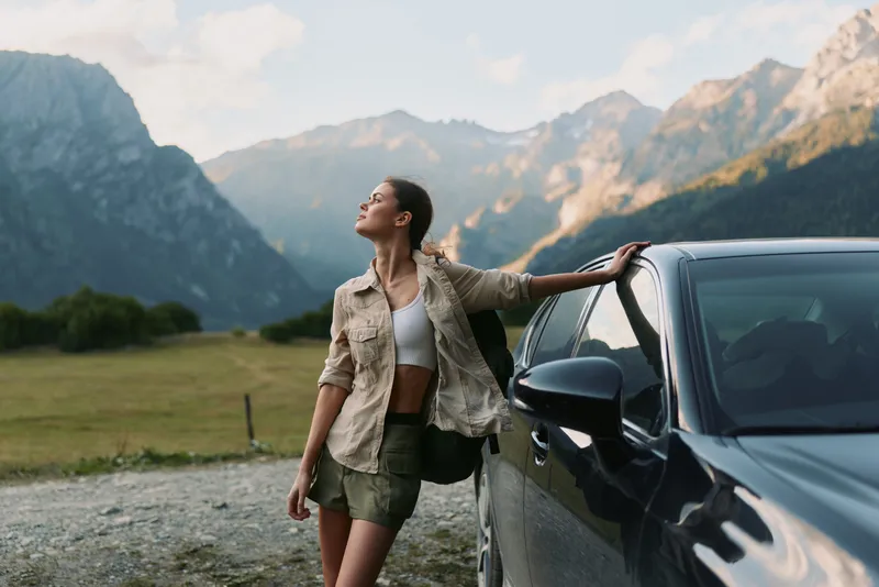 Young woman leans against a car in a mountain valley, gazing upward with a relaxed posture and a sense of freedom under clear skies, sunlight warming her profile.