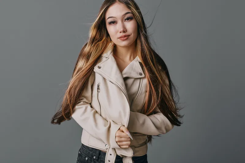 Young woman with long hair wearing beige jacket posing standing on gray background. Female model looking at camera with neutral expression and casual style indoors.