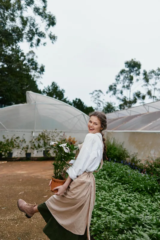 Young woman in a greenhouse, wearing a light blouse and apron, smiling while holding a potted plant perfect for themes of gardening and sustainability