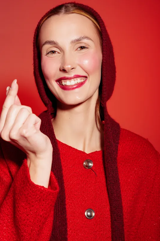 Smiling woman in red sweater and hood makes finger heart gesture on red background. Close-up portrait with bright makeup and joyful expression. Concept of love, happiness, and fashion.
