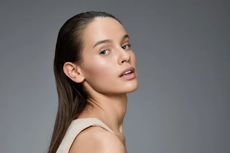 Elegant young model with natural makeup and long hair looking confidently at the camera against a minimalistic grey background, emphasizing beauty and simplicity