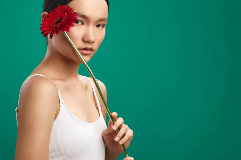 Asian woman in a white tank top holds a red flower near her face against a solid green backdrop, creating a calm, minimalist portrait with soft lighting and serene mood.