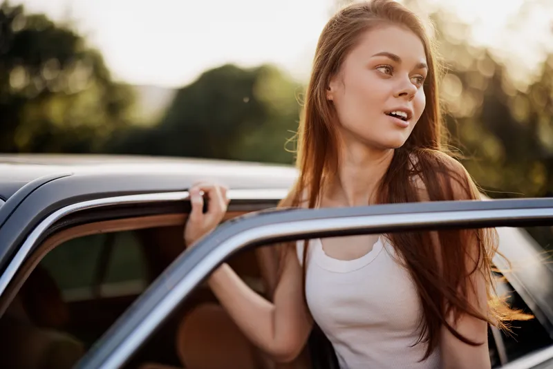 Young woman in casual summer attire enjoying the outdoors, leaning out of a car, radiating confidence and joy amidst a lush green background.