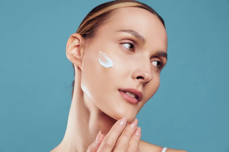Young woman applying face cream on clean skin, smooth complexion, natural beauty, gentle skincare routine, soft lighting, blue background, close-up portrait