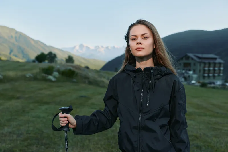 A confident young woman stands outdoors in a windy landscape, holding a trekking pole. She wears a jacket and gazes forward, suggesting adventure, resilience, and a readiness to explore.