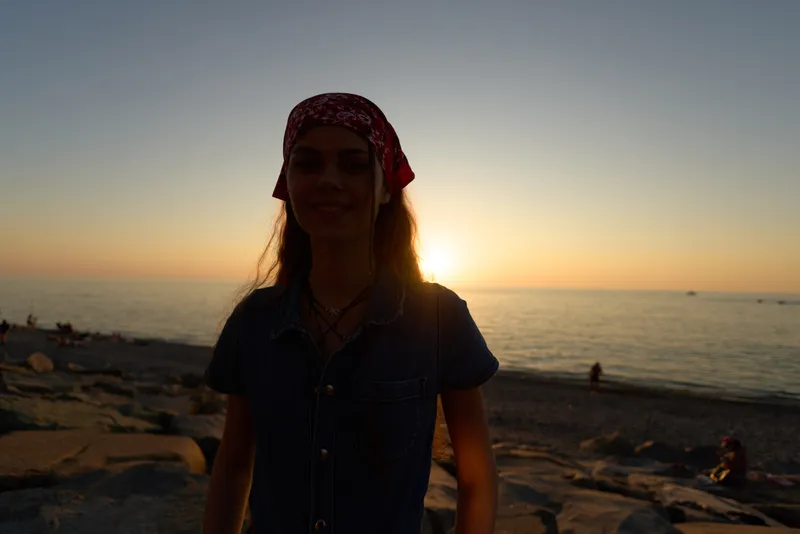 woman standing at the beach during sunset wearing a red headscarf and denim clothing with calm sea and warm sky in background