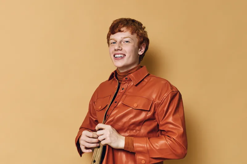 Young man with red hair smiling and wearing orange jacket and turtleneck on beige background. Casual style, happy expression, portrait, confident and cheerful mood in studio.