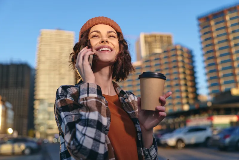 woman coffee phone smile city beanie sunlight morning candid lifestyle portrait of a smiling woman holding coffee and phone in urban golden hour glow, conveying authenticity, mindful living and