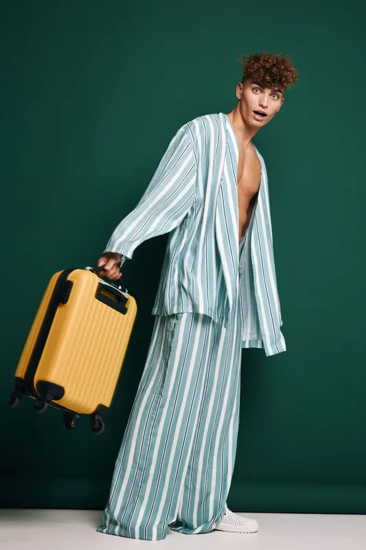 Young man in oversized striped pajamas holding yellow suitcase with accepted luggage bags in playful pose travel concept against dark green background for vacation or airport scenes.