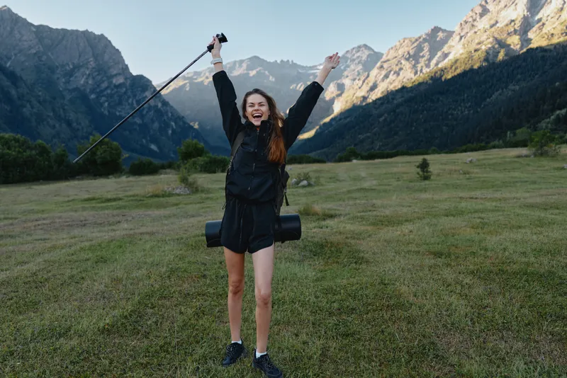 A joyful hiker stands in a wide grassy valley, arms raised in triumph, trekking pole in hand, backpack secured. Rugged mountains rise in the distance beneath a clear sky.