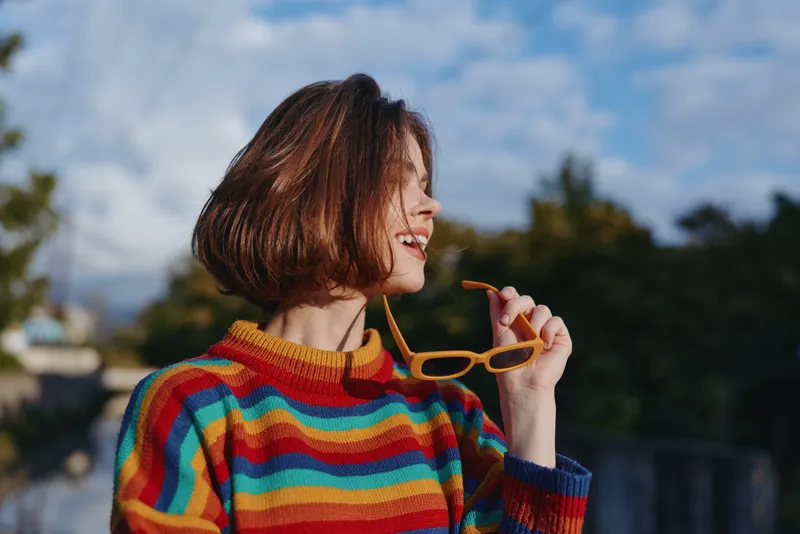 Woman portrait smiling in colorful striped sweater holding sunglasses outdoors, short hair profile, casual fashion, joyful expression with natural light and blue sky background