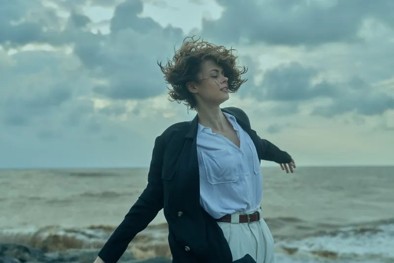 Wind, beach, ocean, movement, coat, shirt frame a windy scene as a woman on the shore stretches arms, faces the sea, and basks in the dramatic sky with rolling waves and misty light