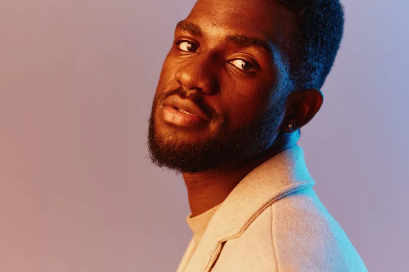 A young Black man with a stylish hairstyle poses confidently in a light gray blazer against a colorful gradient background, exuding charm and sophistication