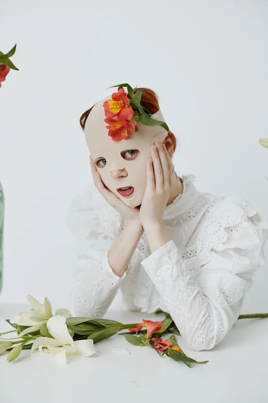 A young model with a creative facial mask and floral headpiece expresses surprise, surrounded by fresh flowers and minimalistic decor