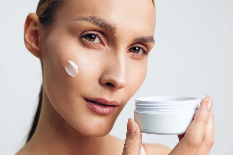 Woman applying white face cream, holding a small jar, close up portrait on light studio background, concept of skincare, beauty, wellness, self care, soft lighting, calm expression, natural glow