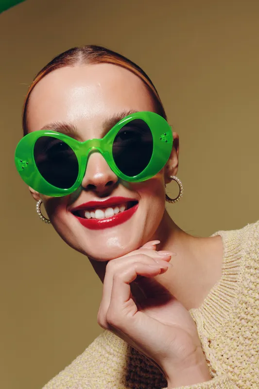 Smiling woman with bright red lipstick wearing large green sunglasses and hoop earrings poses with hand near face. Fashion portrait with warm lighting and beige background. Stylish mood.