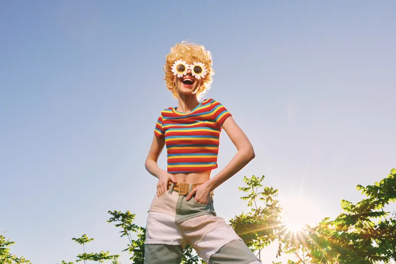 Cheerful woman outdoors, wearing striped crop top and retro sunglasses, sunlit moment, energetic pose, carefree summer mood, vivid colors, natural greenery background, playful spirit