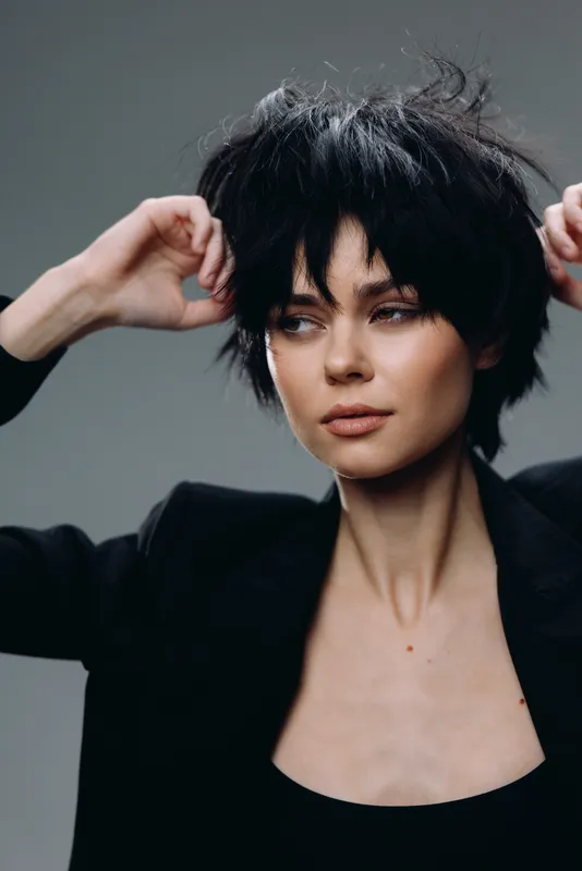 Young woman with short black hair wearing dark clothing posing confidently against a gray background.