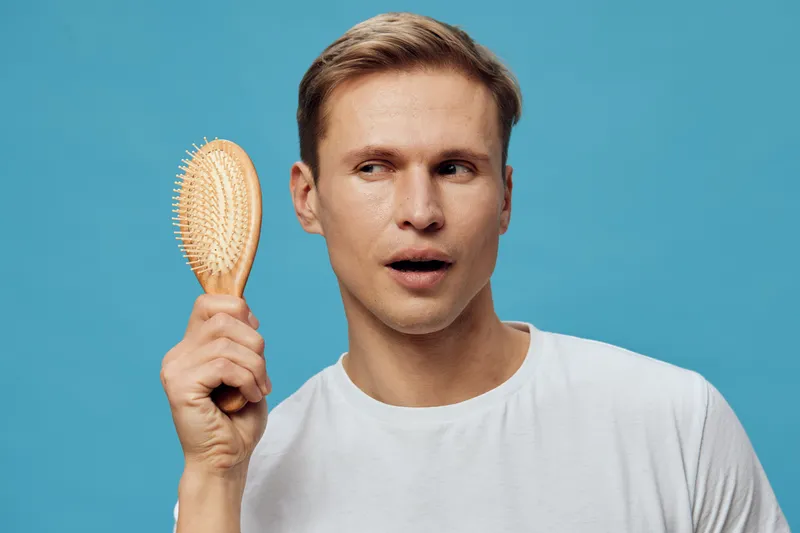 surprised young caucasian man hold hairbrush near ear curious look isolated on blue background casual white shirt mental health and gadgets concept