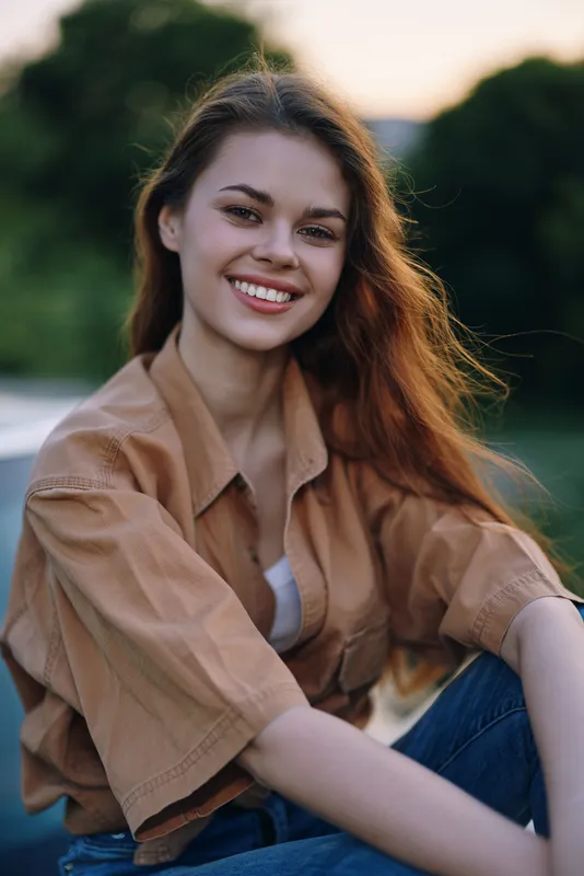 Young woman with long hair smiling in casual outfit against nature background during sunset, expressing joy and positivity.