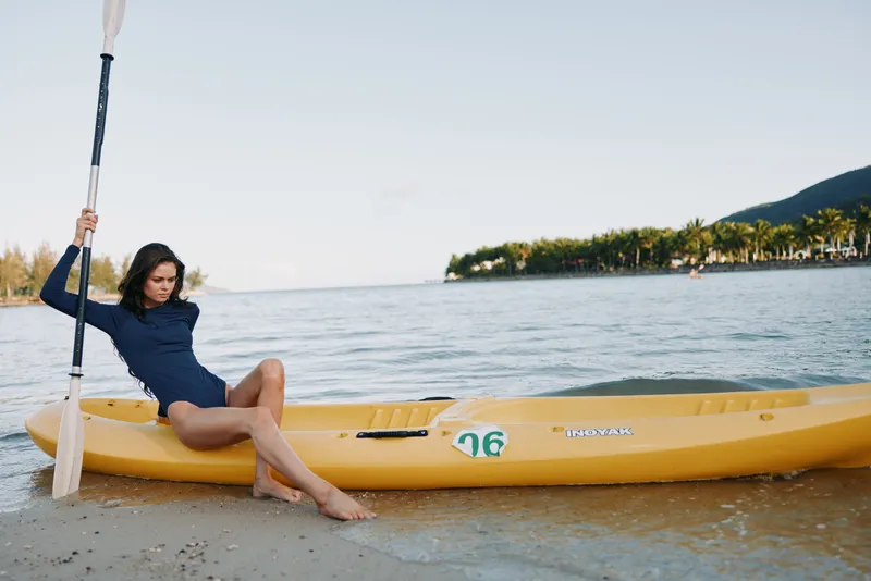 A woman sits on a bright yellow kayak beached on a sandy shore, gripping a paddle, while calm water and distant trees frame a relaxed, sunlit scene.