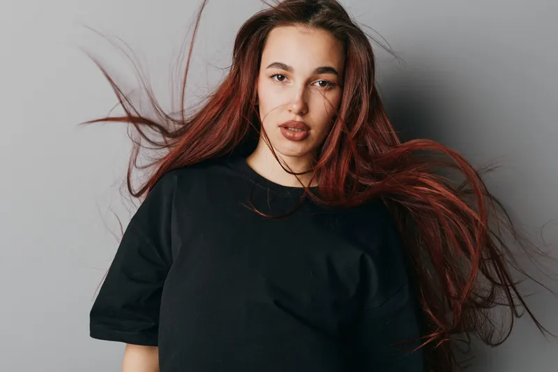 Young woman with long red hair wearing black t shirt posing in studio with hair flowing and neutral expression on gray background for fashion portrait and beauty concept.