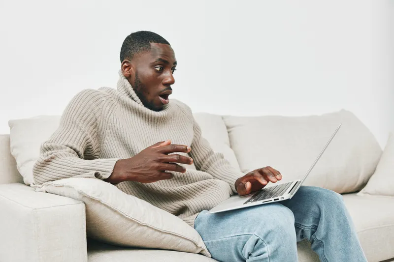 Surprised young Black man in a cozy sweater reacting to shocking news while using a laptop in a modern living room with neutral tones