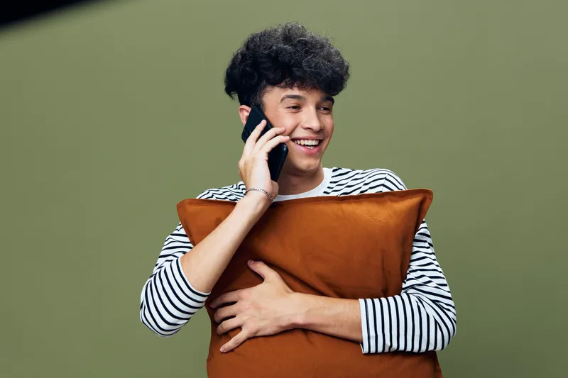 smiling man talking on phone holding brown pillow wearing striped shirt with curly hair on green background, positive emotions and casual style
