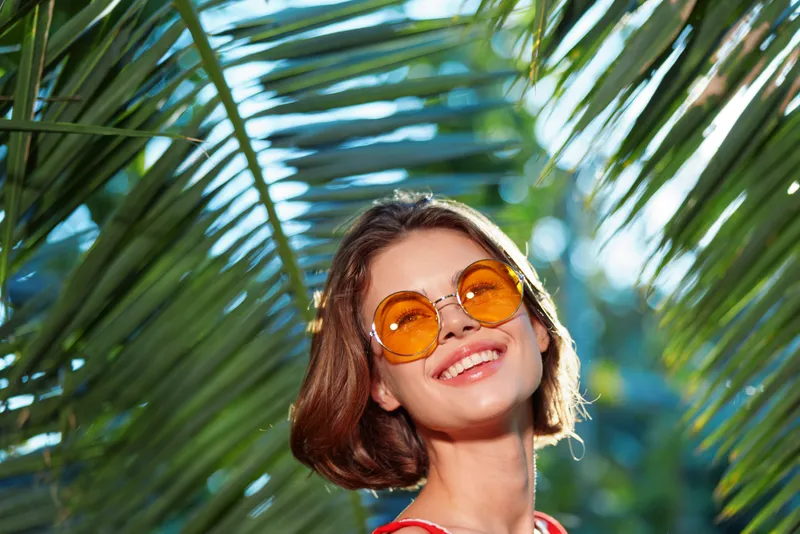 A cheerful woman wearing round orange sunglasses looks up with a bright smile in a sunlit tropical garden, surrounded by lush palm fronds and vibrant light.