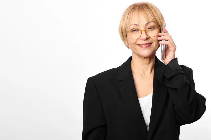 Smiling senior woman with glasses in a studio portrait, wearing a black suit jacket, touching her glasses with one hand, clean white background, calm confidence