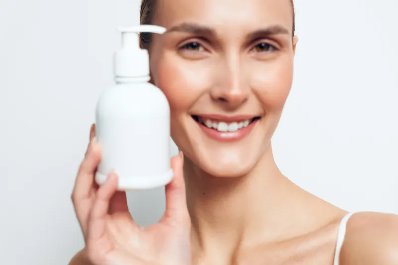A smiling woman with clear skin holds a white plastic pump bottle close to her face. The bright background emphasizes skincare and beauty product concepts for personal care routines.