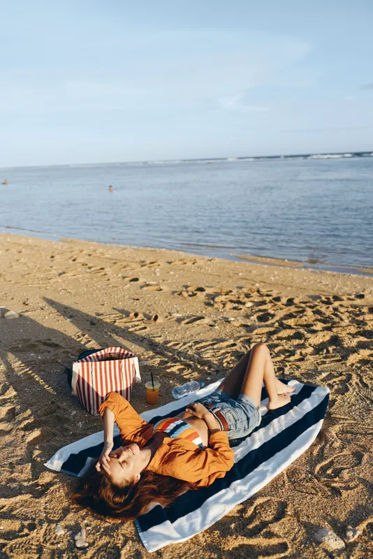 Young woman with long hair relaxes on a striped towel on the sandy beach near the calm sea during a sunny day.
