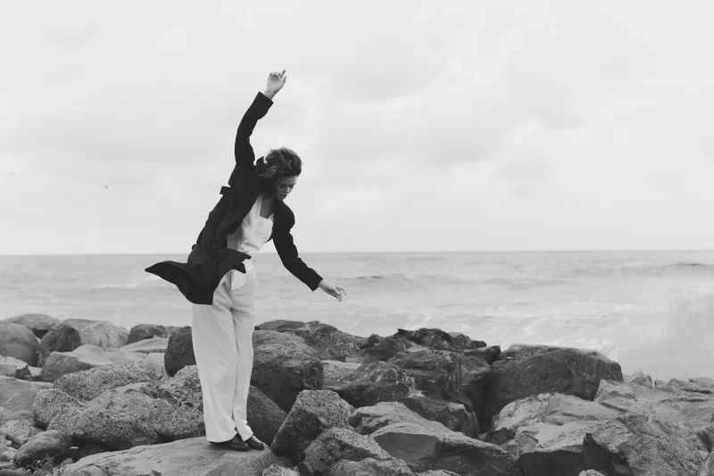 Dance on a rocky shore by the sea, a figure in a dark jacket lifts an arm in motion against a cloudy sky, captured in stark blackandwhite for a dramatic, timeless scene