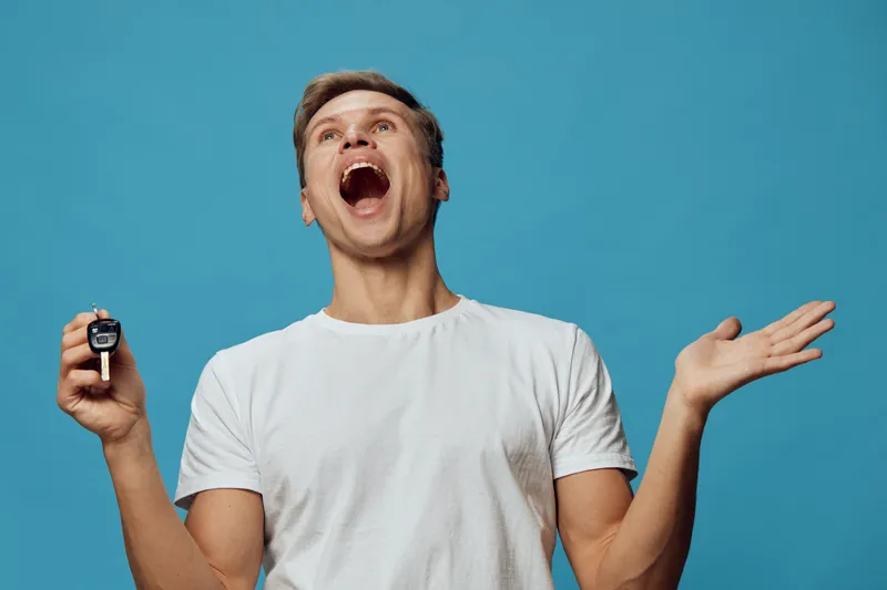 Joyful man celebrating happiness, holding car key, expressing excitement in casual white t-shirt against blue background, lifestyle and achievement concept.