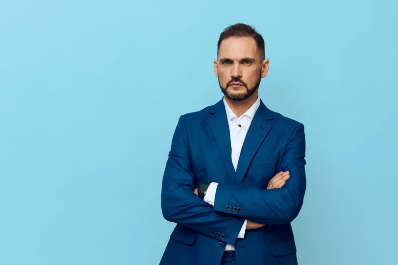 A confident business professional stands against a solid blue backdrop, projecting calm authority and focused demeanor. The isolated color emphasizes leadership mood and purposeful presence in a