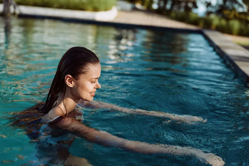A smiling young woman with wet hair enjoys swimming in an outdoor pool on a sunny day. The scene captures a relaxed lifestyle and leisure activity in a natural setting.