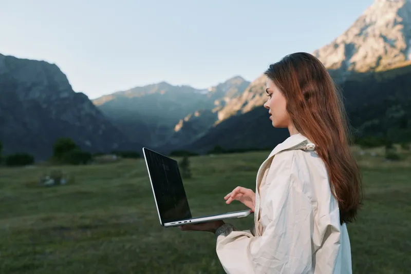 A focused woman uses a laptop outdoors in a scenic mountainous field. She types intently in a light jacket, with sunlit peaks in the background, projecting calm, productive energy.