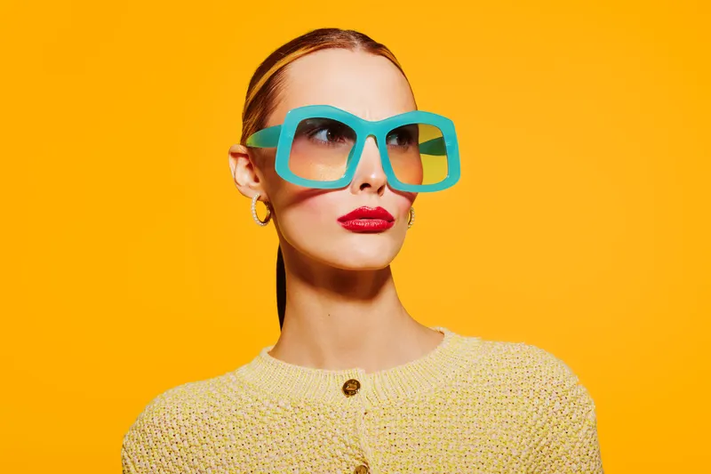 Stylish woman with large turquoise sunglasses and red lipstick poses against bright yellow background. Fashion portrait with retro vibe and bold color contrast for beauty and style concept.