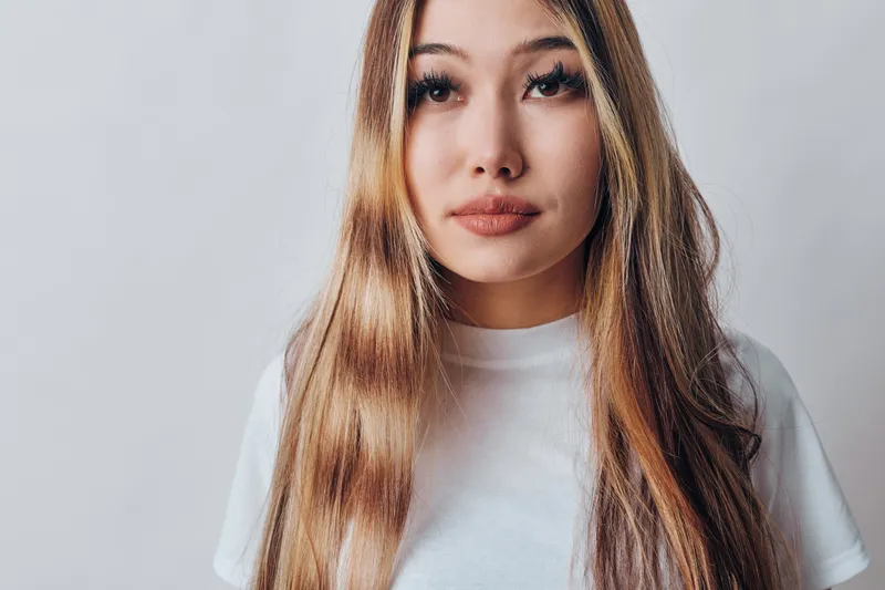 Young woman with long hair and natural makeup wearing white t shirt standing against plain light background with calm facial expression and direct gaze.