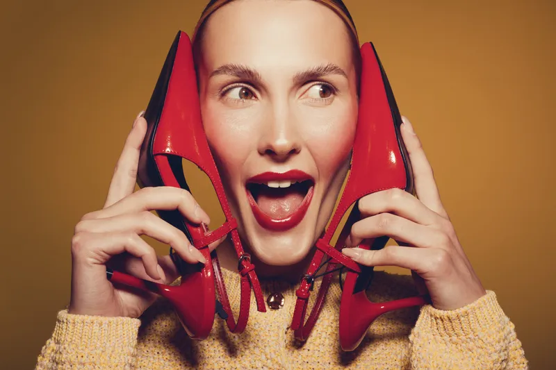 Young woman with red lipstick holds red high heels near her face, smiling with open mouth on yellow background. Concept of fashion, style, and playful mood in studio lighting.