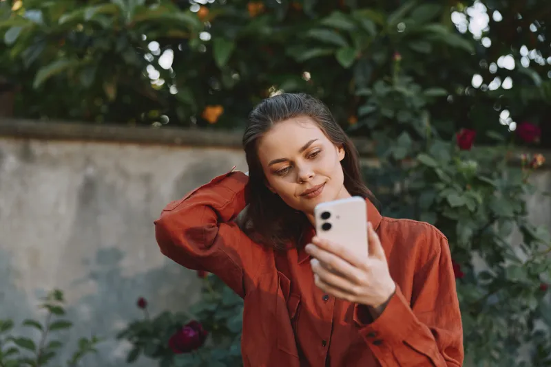 Smiling woman in an orange sweater taking a selfie outdoors, garden backdrop, lifestyle moment, casual vibe, natural light, warm and relaxed, everyday beauty